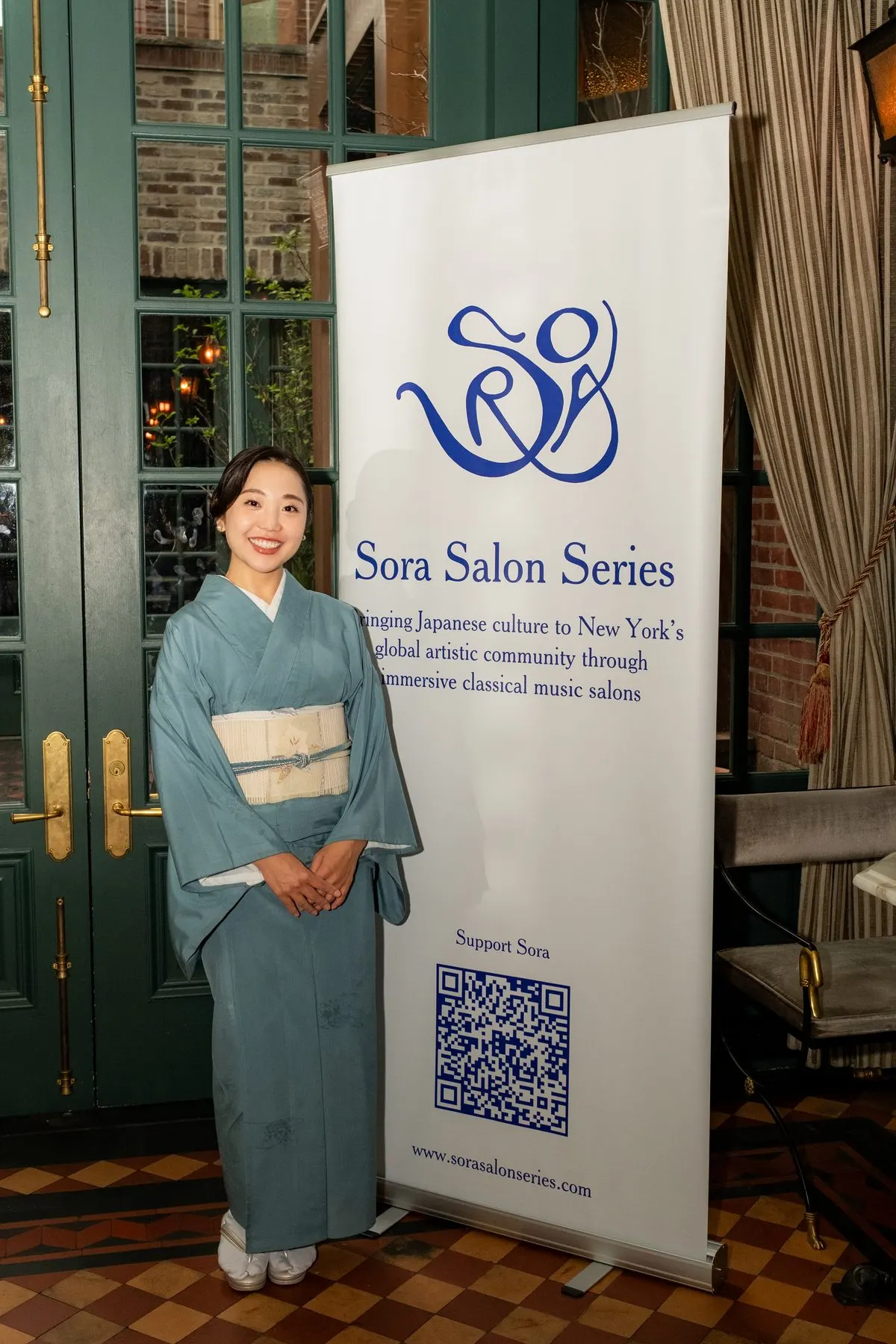 A host in light blue kimono beside the Sora banner at the entrance