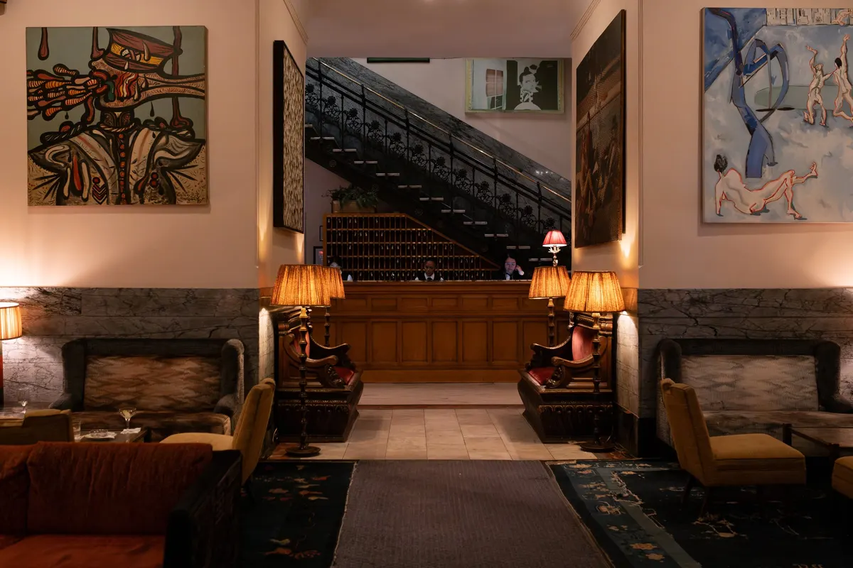 The lobby of The Hotel Chelsea: the iron staircase over the front desk, lamps lit, a painting on each side